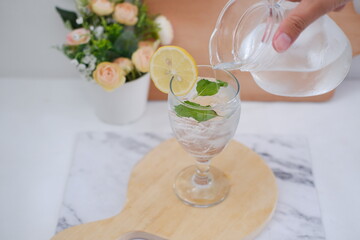 Pouring water into glass with mint and lemon on white marble table