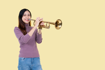 A talented Asian woman is playing a trumpet while standing against an isolated yellow background.