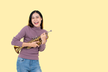 A joyful Asian woman holding a trumpet, stands against an isolated yellow background.