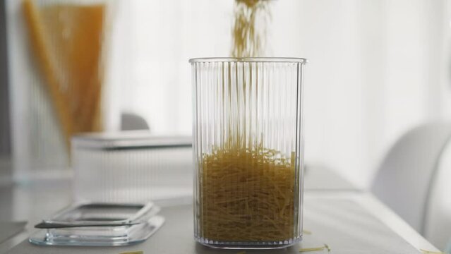 Broken pasta falls into tall plastic container on table. Person pours raw product into packing for storage in light kitchen. Maintaining order in house