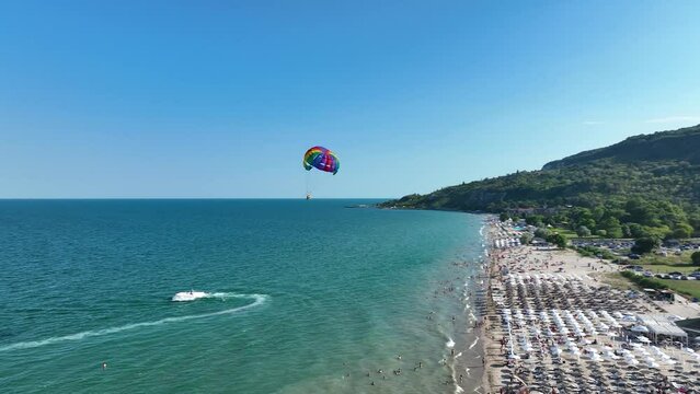 Aerial view of a motor speed boat towing a parachute with people. The parachute rises high above the water against the background of the sea, hotels and beaches. Summer vacation shot