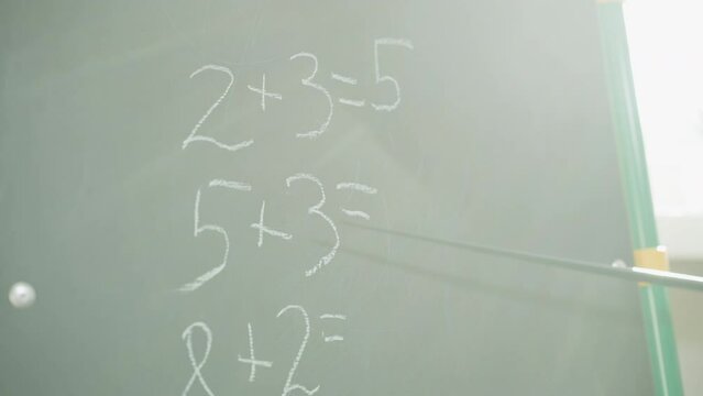Tutor shows unsolved maths task written on blackboard with pointer in classroom closeup. Additional mathematical problems for elementary school on blackboard