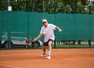 An elderly man playing tennis is hitting a forehand. The moving at camera. Open ground.