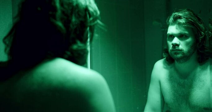 Long haired, bearded naked man looking at himself in the mirror at the bathroom. Green light