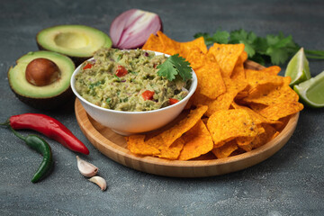 White bowl of traditional Mexican guacamole with nachos on grey concrete background. Tortilla chips with guacamole sauce dip and ingredients: avocado, cilantro, onion, hot pepper and lime