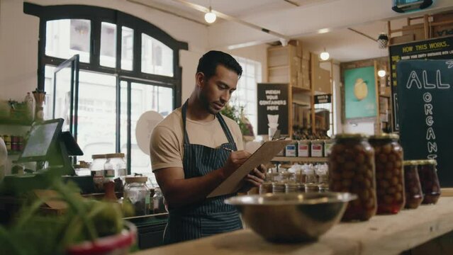 Young multiracial man counting stock of healthy foods while working as greengrocer in greengrocer's shop