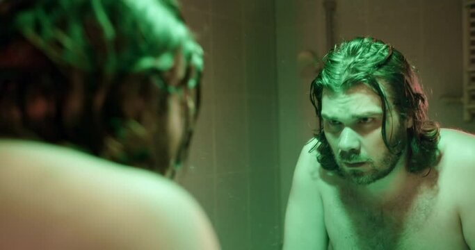 A serious man with long hair looks at himself in the mirror, looking him self in the eye. Sets himself up to be confident, focused. Make affirmation. Man after bath, depressive light  