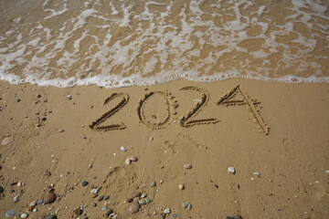 New Year 2024, lettering on the beach with wave and clear blue sea. Numbers 2024 year on the sea shore, message handwritten in the golden sand on beautiful beach background. New Years concept.