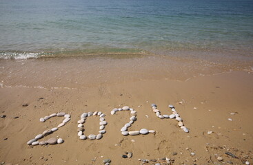 New Year 2024, lettering on the beach with wave and clear blue sea. Numbers 2024 year on the sea shore, message handwritten in the golden sand on beautiful beach background. New Years concept.