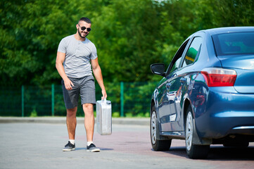 Happy man got full canister of fuel and going to refuel car tank. Smiling male finally found petrol station and fill canister with gasoline. Male going to blue car with cistern in hand.