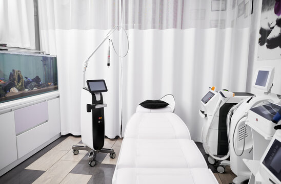 Interior of beautician cabinet with modern cosmetology equipment for skincare rejuvenation treatment and salon chair. Radiofrequency microneedling device, patient daybed and laser machine in clinic.