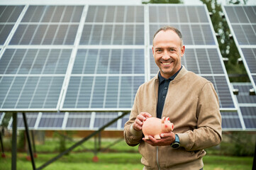 Man happy about his investments in alternative energy. Enthusiastic investor glad about earnings he got. Man with piggy bank in hands smiling to camera.