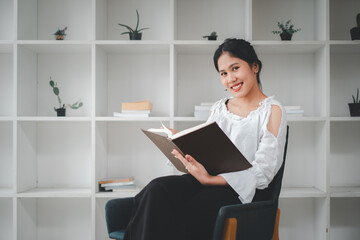 Side view of asian woman sitting armchair with open book and reading in living room.