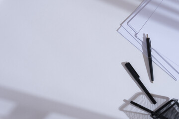 Clean minimalist white office desk with shadow, pens, pen holder, and sheets of white paper
