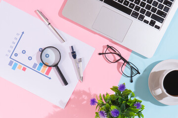 Flowering plant and coffee cup on office desk with laptop, pink and blue background
