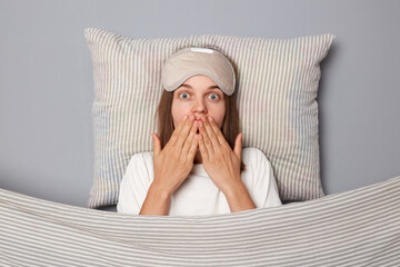 Shocked scared woman in white T-shirt and sleeping eye mask lie in bed on pillow under blanket...