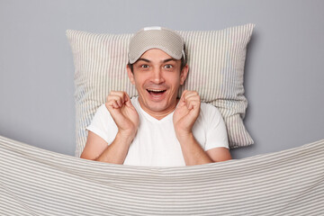 Surprised smiling man with resting on white pillow under blanket wearing t-shirt isolated over grey...