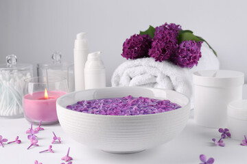 Bowl of water with lilac flowers, spa products and burning candle on white table