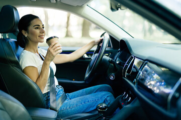 Happy young woman with coffee to go driving her car. Side view of woman driving car with coffee to...