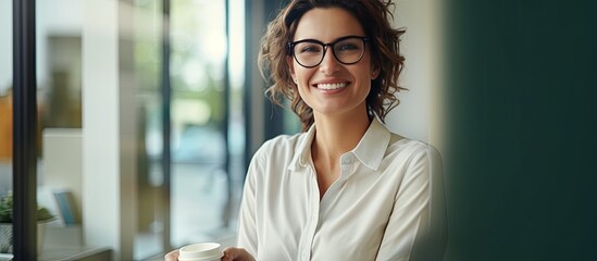 A young caucasian woman confidently stands by a window wearing a shirt smiling happily while holding a coffee cup