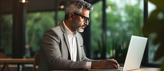 Mature CEO of Indian or Latin origin using laptop in contemporary office analyzing online data and devising tech strategy