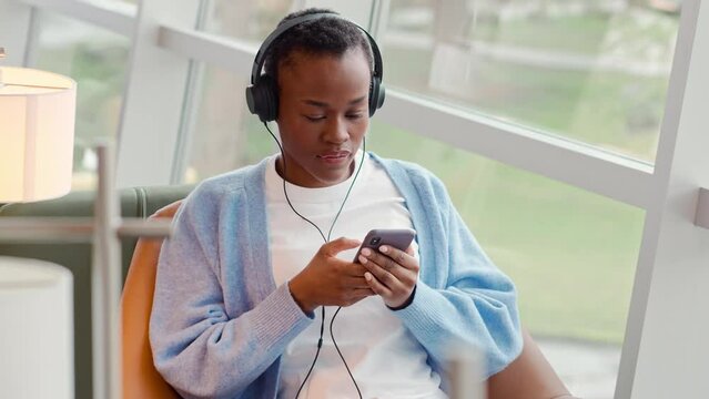 African Black teenage girl university or college student refugee sitting in chair in university campus, coworking office space or airport lounge, wearing headphones looking away at window.