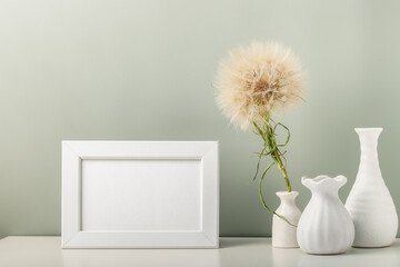 An empty photo frame and three white vases and a dandelion on the table.