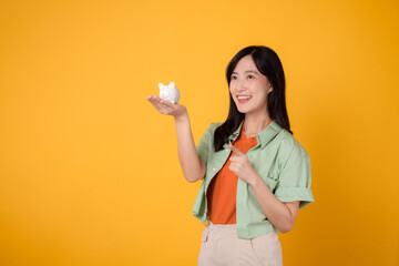 Fototapeta na wymiar cheerful young Asian woman in her 30s, donning orange shirt and green jumper, displaying piggy bank while pointing finger to free copy space on yellow background. Financial money concept.
