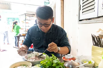 Poster A man using chopsticks and spoon eating traditional Pho Bo vietnamese soup with beef and rice noodles on a metal table, real scene in local restaurant © CravenA
