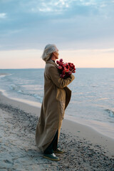 Lonely caucasian middle-aged woman with a bouquet of flowers on seashore in evening, vertical angle