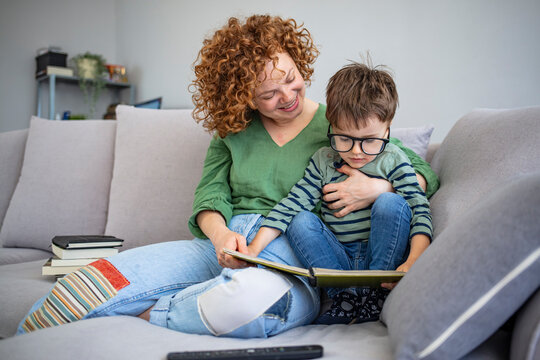 Little boy and his mother are reading a book together in the living room of their home. Mom and little kid son read book.  Mother sitting in an armchair reading a book with her three year old son