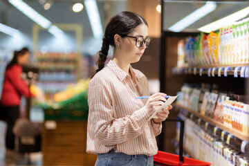 Inspection of grocery store. Portrait of young serious thoughtful caucasian woman wearing eyeglasses writing in document. Concept of shopping in supermarket and consumerism - obrazy, fototapety, plakaty