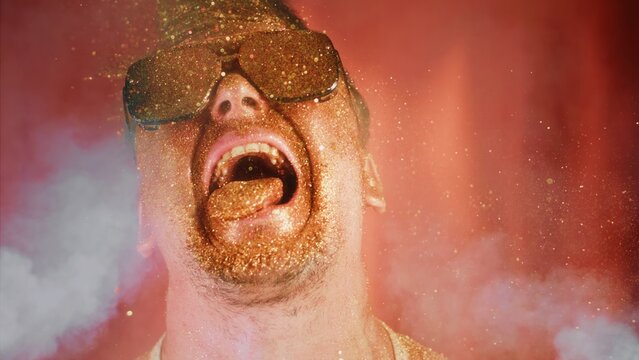 Crazy man sprinkles himself with sparkle particles. Fun video, funny face, tongue out. Happy joy portrait male person in sunglasses pouring on face and beard gold glitter.Funny crazy man in gold dust
