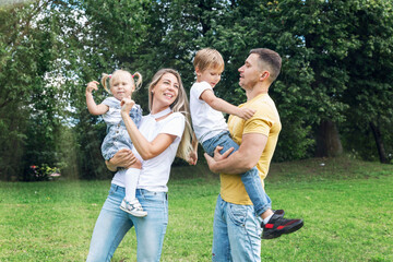 Fototapeta premium Happy family with children in the park on a sunny day. Mom, dad and little daughter and son in jeans and white t-shirts laugh and hug. Love and tenderness.