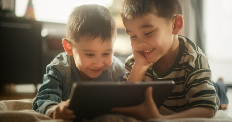 Portrait of Two Male Asian Kids Using a Digital Tablet During Weekend in their Room. Two Cute Brothers Watching Cartoons Together, Focusing on the Screen, Sharing the Gadget and Enjoying their Time