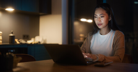 Beautiful Authentic Asian Woman Sitting at a Table in a Cozy Kitchen and Typing On Laptop Computer at Home at Night. Female Smiling and Chatting on Social Media, Doing Online Shopping, Checking News.