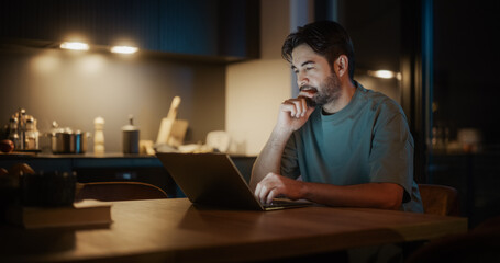 Handsome Authentic Latin Man Sitting at a Table in a Cozy Kitchen and Using Laptop Computer at Home at Night. Male Adult Thinking While Browsing the Internet, Checking Videos on Social Networks.