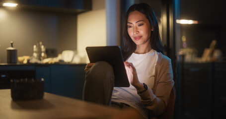 Beautiful Authentic Asian Woman Sitting at a Table in Cozy Kitchen and Using Digital Tablet at Home...
