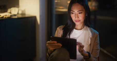 Beautiful Authentic Asian Woman Sitting at a Table in Cozy Kitchen and Using Digital Tablet at Home at Night. Female Smiling and Checking Social Media, Doing Online Shopping, Watching Internet Videos
