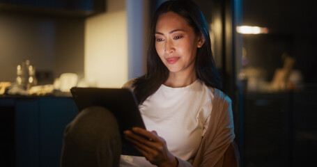Beautiful Authentic Asian Woman Sitting at a Table in Cozy Kitchen and Using Digital Tablet at Home at Night. Female Smiling and Chatting on Social Media, Doing Online Shopping, Browsing the Internet