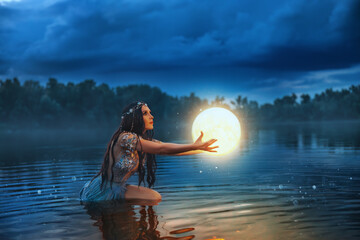 Art photo magic fantasy woman holding moon universe planet ball in hands raises to dark night sky. Mystic witch Girl fairy elf river nymph posing sitting in water lake summer nature. Lady sexy queen