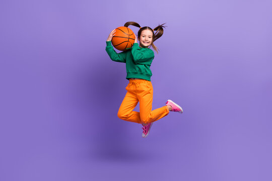 Full length photo of excited funky small girl wear green sweatshirt jumping high rising basketball ball isolated violet color background
