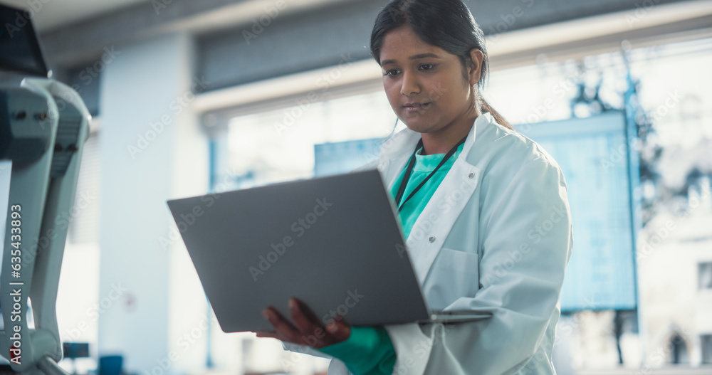 Fototapete Young Indian Female Engineer Testing Futuristic Programmable Robot Animal in a Factory Development Workshop. Professional Researcher in a Lab Coat Developing AI Canine Prototype, Using Laptop