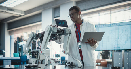 Naklejka na ściany i meble Young African American Male Engineer Testing Industrial Programmable Robot Animal in a Factory Development Workshop. Black Researcher in a Lab Coat Developing AI Pet Prototype, Using Laptop