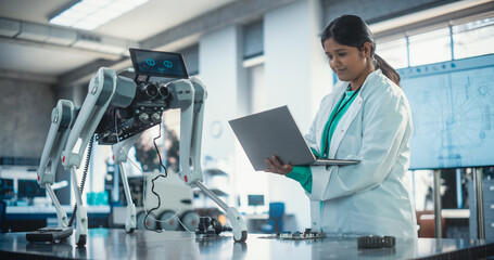 Young Indian Female Engineer Testing Industrial Programmable Robot Animal in a Factory Development Workshop. Professional Researcher in a Lab Coat Developing AI Canine Prototype, Using Laptop