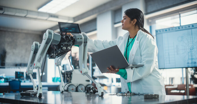 Young Indian Female Engineer Testing Industrial Programmable Robot Animal in a Factory Development Workshop. Professional Researcher in a Lab Coat Developing AI Canine Prototype, Using Laptop Computer