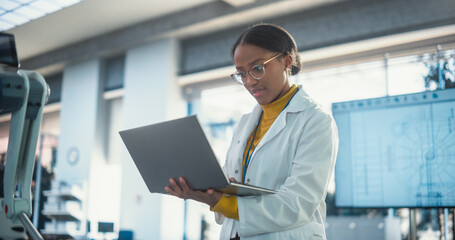 Portrait of Young Black Female Specialist in Lab Coat Using Laptop to Test an AI Robotic Prototype. Professional, Successful Woman Working as an Engineer in Modern High Tech Company Startup