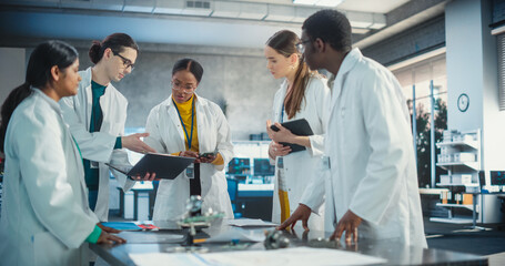 Fototapeta premium Engineers In Lab Coats Using Laptop and Tablet, Brainstorming Ideas For Electronic Components. Diverse Team of Industrial Robotics Specialists Gathered Around a Table to Discuss a Blueprint.