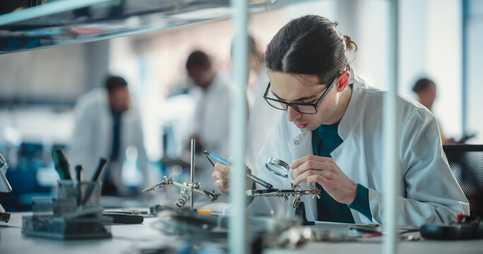 Diverse Young Group of Engineers Working in a Startup Lab. A Team of Professionals Discuss Technological Project Strategy Using Laptop Computer While Male Specialist is Soldering a Circuit Board