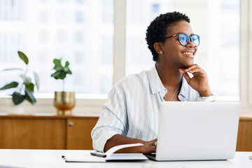 Portrait of dreamy african american businesswoman sitting at the desk in front of the laptop and thinking, female office employee in glasses looking away with a light smile, satisfied with work done